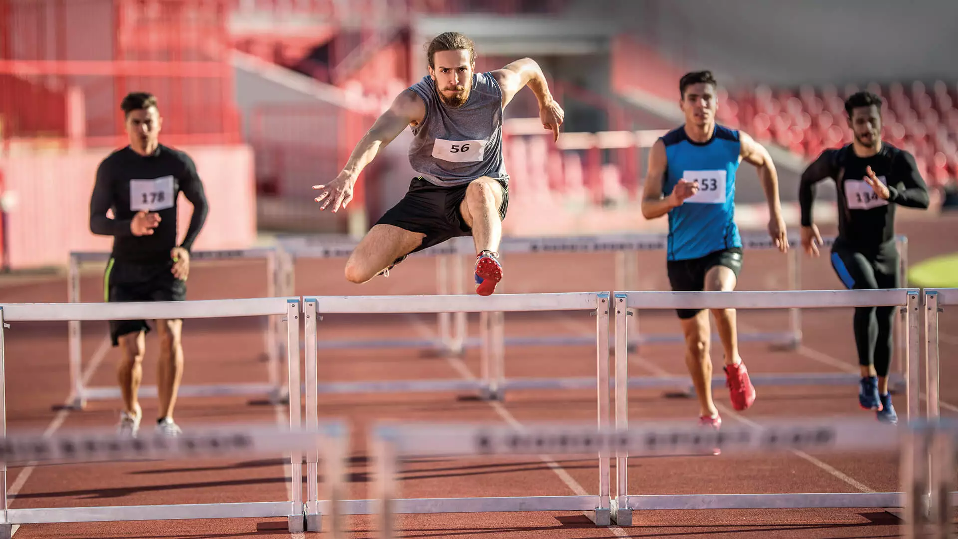 Das Bild zeigt mehrere Athleten, die an einem Hürdenlauf teilnehmen. Im Vordergrund sieht man einen Läufer, der gerade über eine Hürde springt, während weitere Teilnehmer im Hintergrund zu sehen sind, die ebenfalls über Hürden springen oder sich darauf vorbereiten. Die Szene spielt sich auf einer Leichtathletikbahn ab, die mit typischen roten Belag versehen ist. Die Athleten tragen sportliche Kleidung und Startnummern, und es herrscht eine dynamische, wettkampforientierte Atmosphäre.