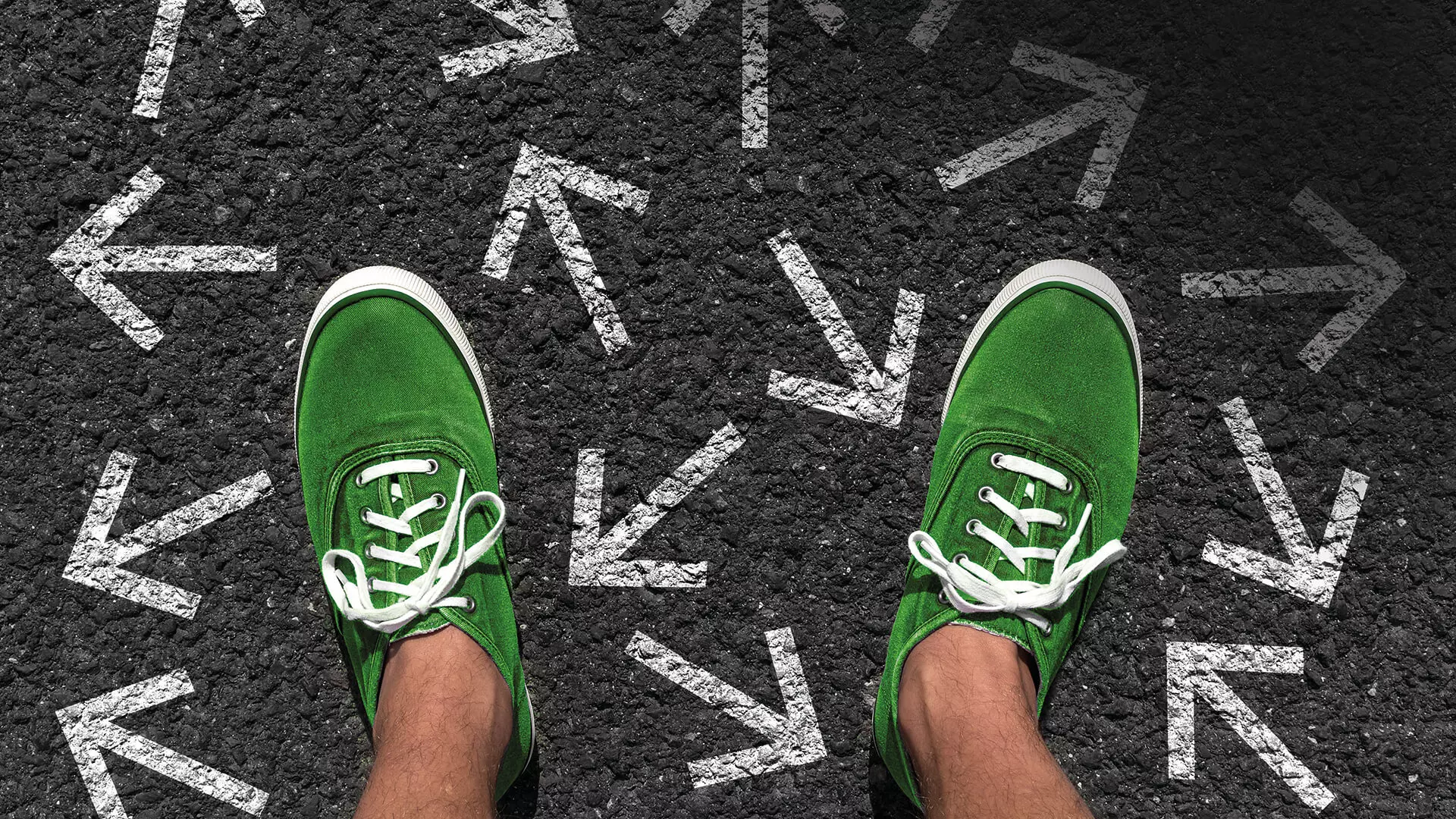 Das Bild zeigt ein Paar grüne Schuhe, die auf einer asphaltierten Straße stehen, auf der viele weiße Pfeile in verschiedene Richtungen gemalt sind.