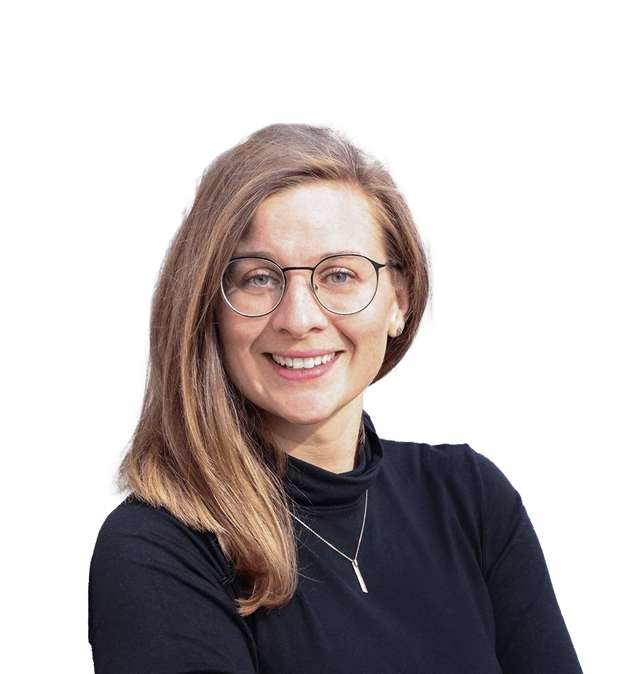 Eine Frau mit schulterlangem, hellbraunem Haar lächelt direkt in die Kamera. Sie trägt eine runde Brille und ein schwarzes Oberteil mit einem hohen Kragen. Um den Hals trägt sie eine feine Halskette mit einem kleinen Anhänger. Ihre Augen sind blau, und ihr Gesichtsausdruck ist freundlich und herzlich. Der Hintergrund ist weiß, wodurch der Fokus vollständig auf der Frau liegt.