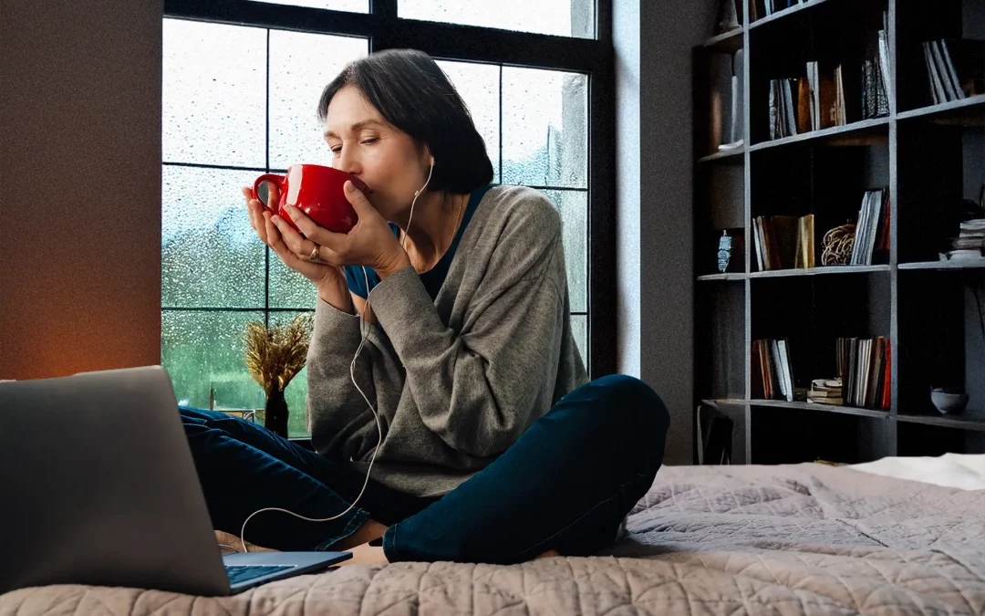 Frau am verregneten Fenster trinkt aus roter Tasse und sitzt mit Laptop auf dem Bett – gemütliche Online-Session im Homeoffice.
